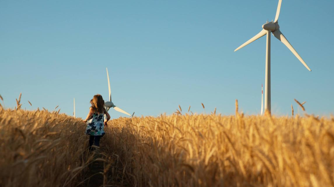 Girl in floral dress is running in corn field with wind turbines