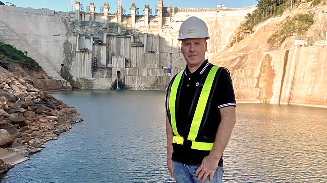 Project manager standing on river's edge with power dam in background