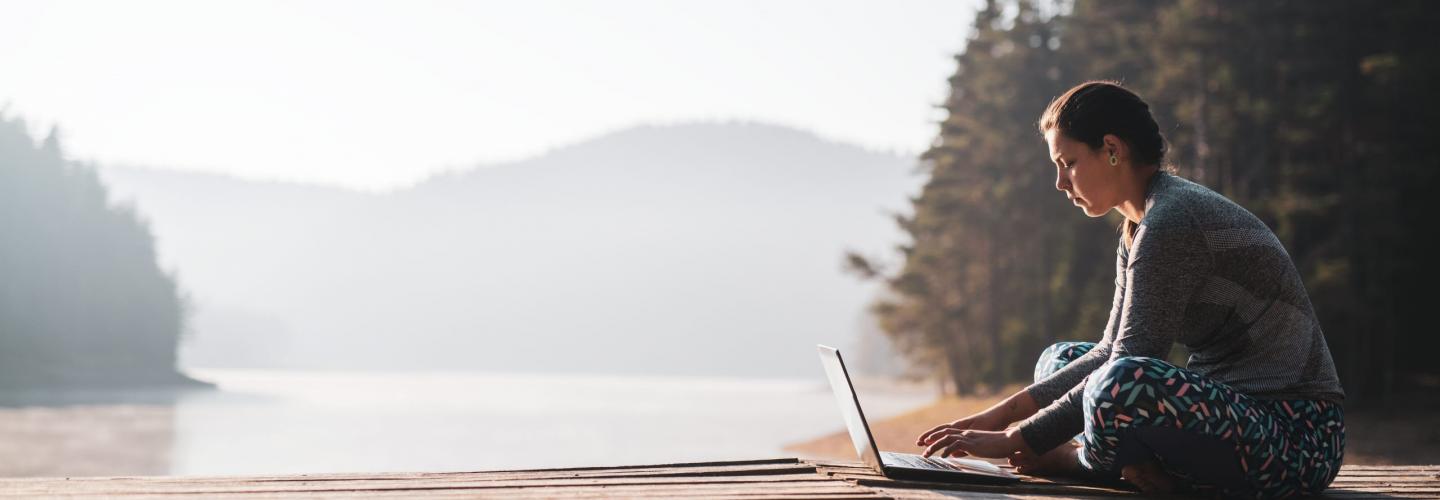Woman using laptop in nature, she sitting on wooden pier around beautiful mountain lake and using laptop.