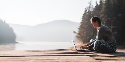 Woman using laptop in nature, she sitting on wooden pier around beautiful mountain lake and using laptop.