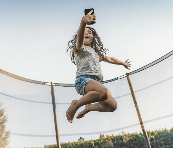 A girl jumping trampoline and taking a selfie with her mobile phone.