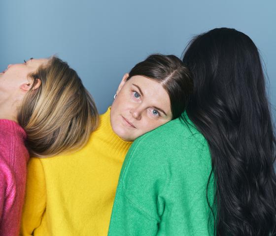Three women standing close to each other, one in a pink sweater, one in yellow and one in green
