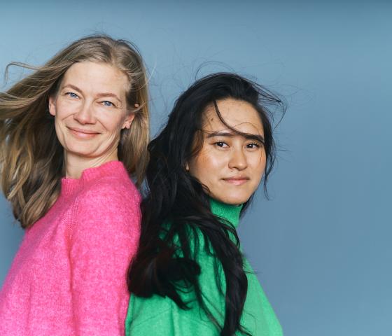 two women standing back-to-back, smiling, one with a pink shirt and one with a green.